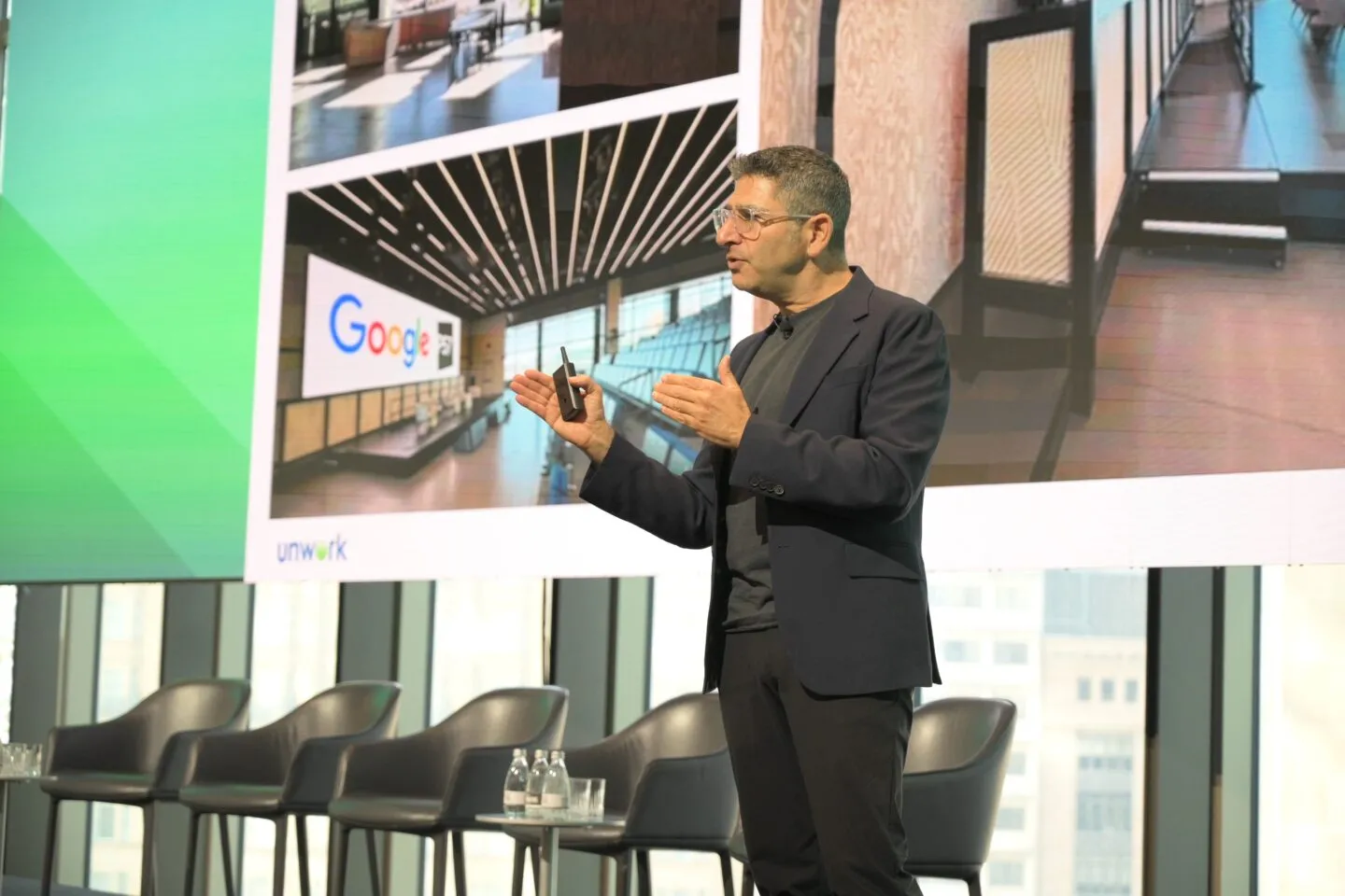 Philip Ross speaking on stage engaging the audience with a talk white 4 balack empty chairs behind and a screen showing workplace images and a subtle Google logo in the background