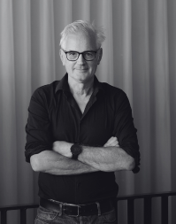 A white-haired man wearing glasses and a dark shirt standing with his arms folded