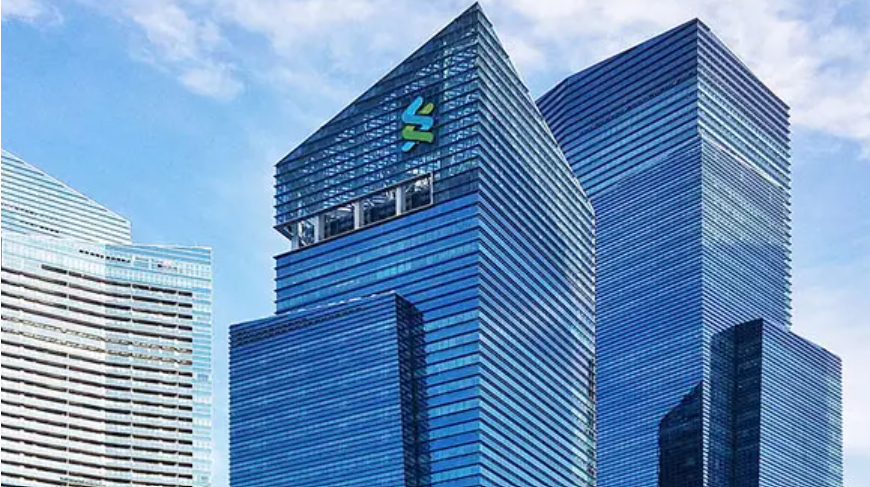 External view of the top of a glass building with a Standard Chartered logo on the top in front of a blue sky