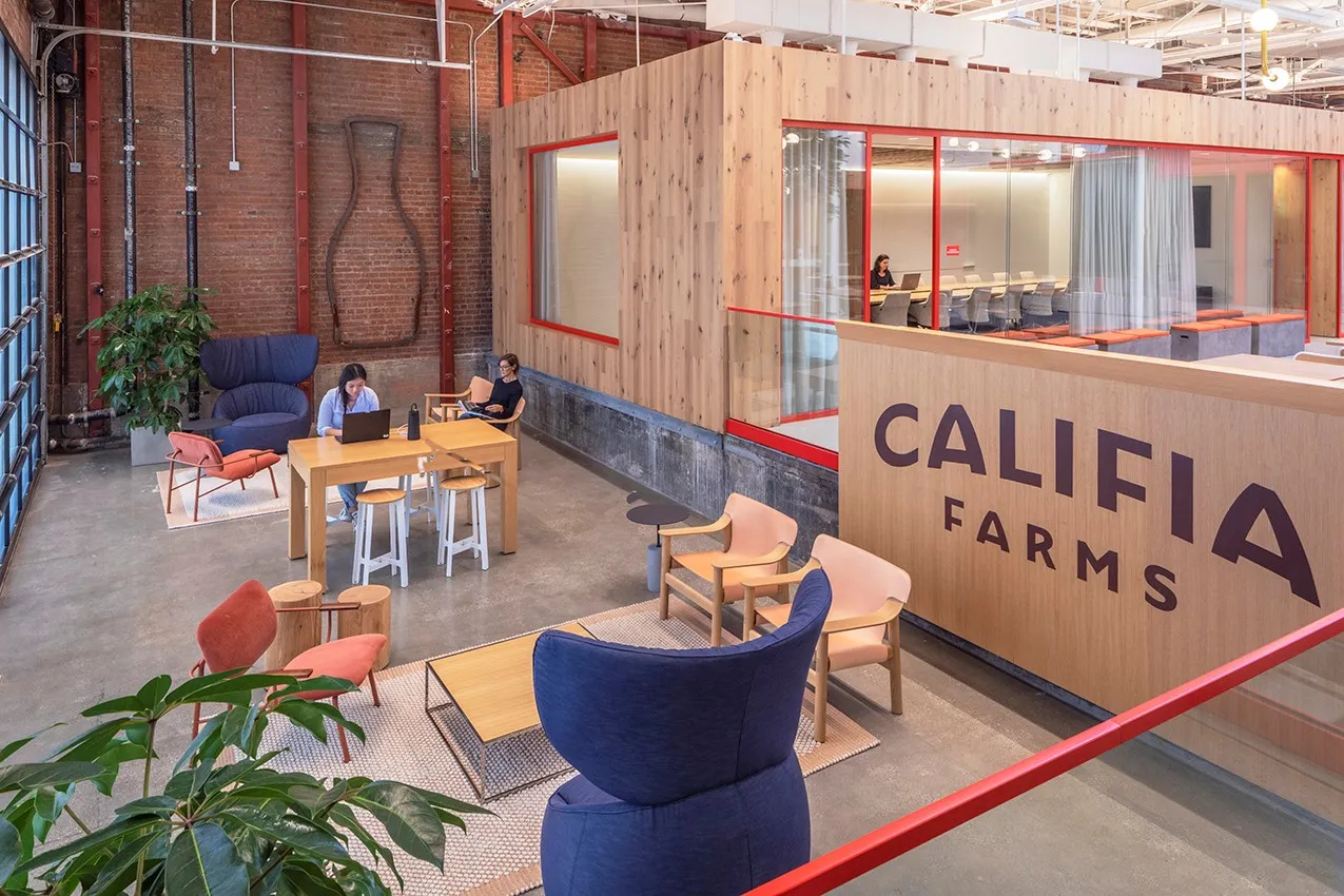 Califia Farms’ Los Angeles headquarters. A red brick space with wooden structures to house meeting rooms decorated with soft seating and wooden worktables and plants.
