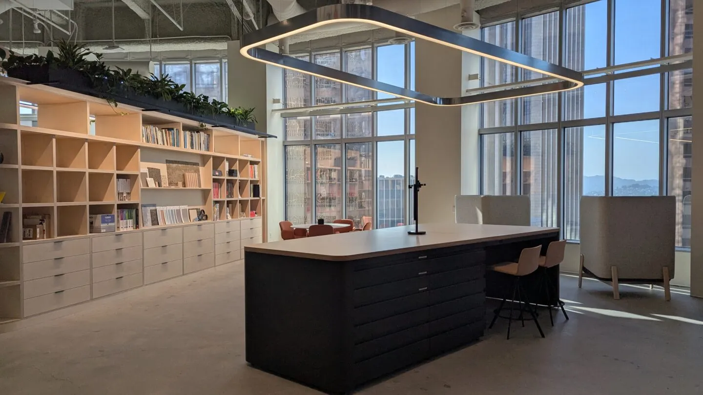 Light open workspace with a central table and overhead circular light fitting, large glass windws with large chairs looking outside and a wall of bookshelves n light wood filled neatly with books and boxes