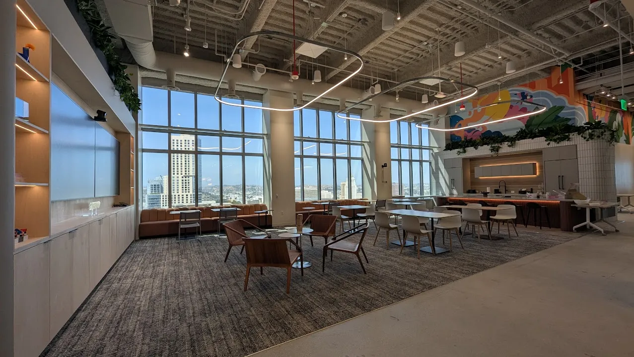 open plan cafe style space with an open kitchen and colourful artowkr above, several small tables and chairs, large slim circule light fittings above, large windows in the background overlooking the city view and a large screen on the left wall with plants above