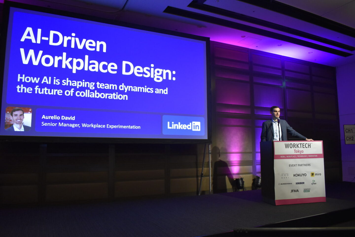 A speaker on stage behind a podium with a large LCD screen in dark room with pink uplighters