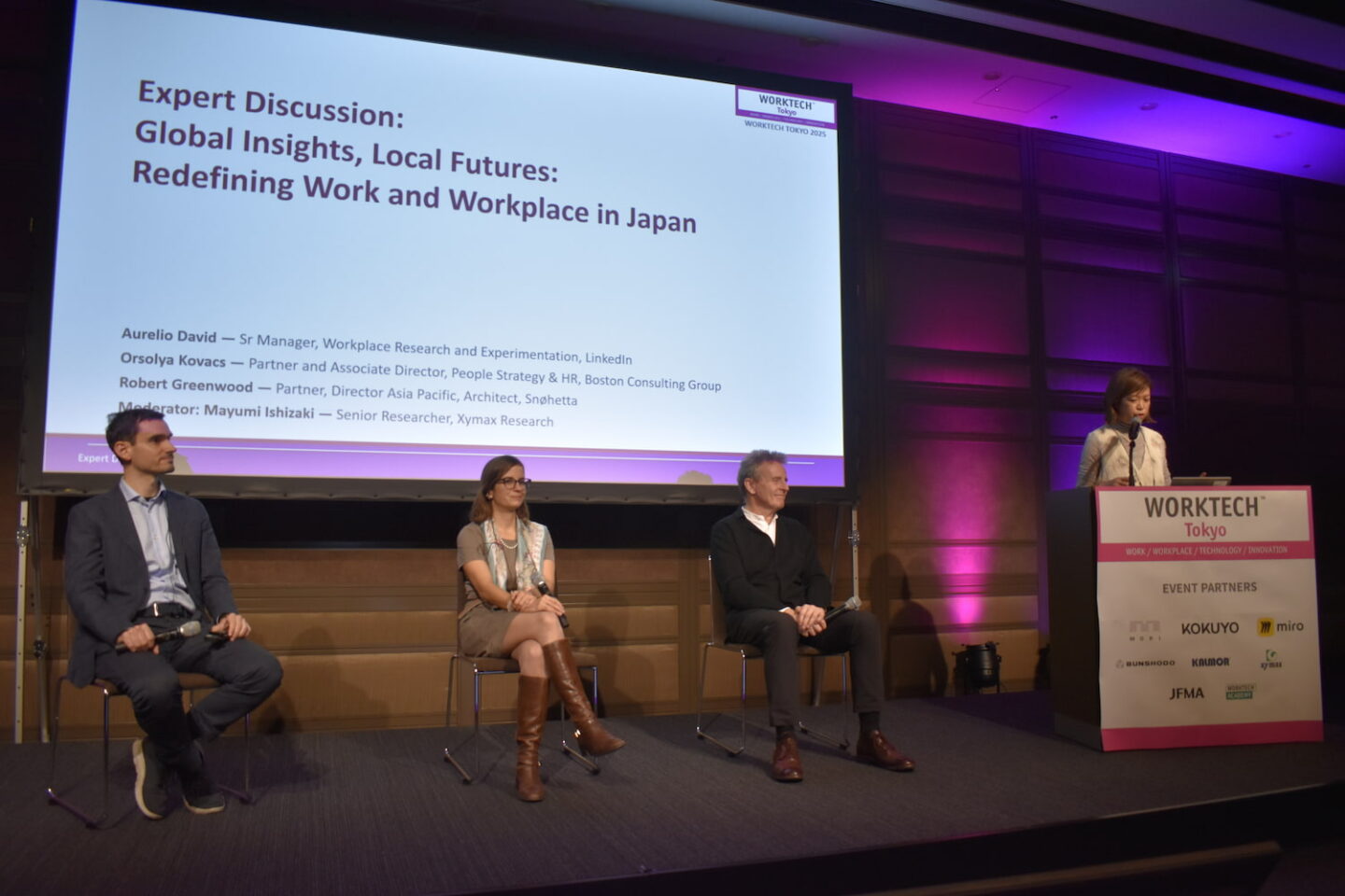 A panel of speakers sat on stage with a moderator behind a podium with a large LCD screen in dark room with pink uplighters